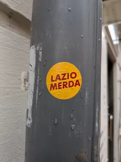 Street sticker A circular, bright yellow sticker is prominently displayed on a dark grey, possibly metallic, drainpipe. The sticker features bold, capitalized red text that reads 'LAZIO MERDA' arranged in two lines, with 'LAZIO' above 'MERDA'. The sticker shows visible signs of wear, including creases and wrinkles, suggesting it is a physical adhesive. Several water droplets are present on both the sticker's surface and the surrounding pipe, indicating an outdoor or damp environment.