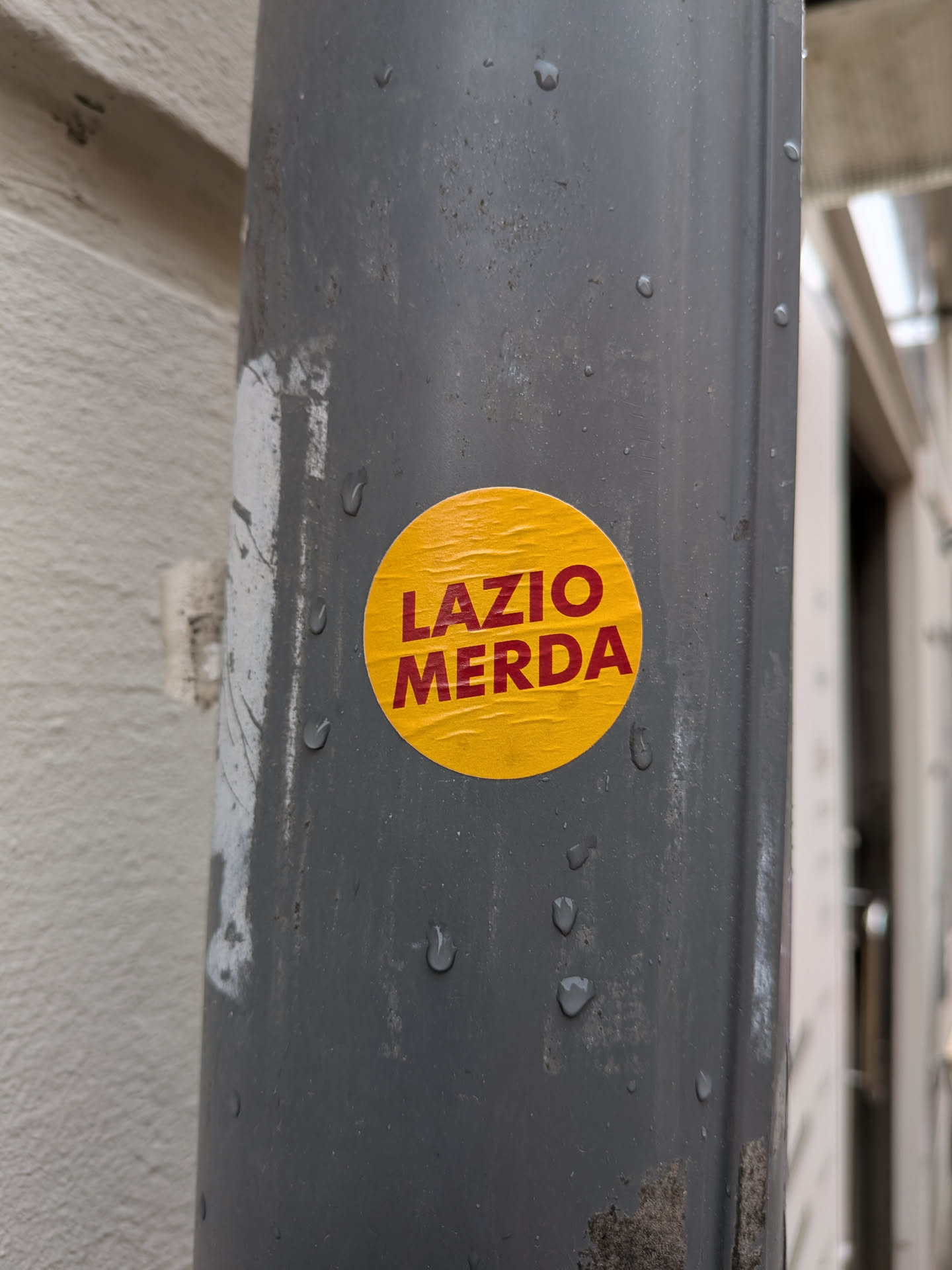 A circular, bright yellow sticker is prominently displayed on a dark grey, possibly metallic, drainpipe. The sticker features bold, capitalized red text that reads 'LAZIO MERDA' arranged in two lines, with 'LAZIO' above 'MERDA'. The sticker shows visible signs of wear, including creases and wrinkles, suggesting it is a physical adhesive. Several water droplets are present on both the sticker's surface and the surrounding pipe, indicating an outdoor or damp environment.