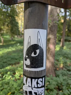 Street sticker The sticker is predominantly white with a black illustration. It depicts the head of a dog or a similar canine creature. The face is filled with a textured black pattern, resembling a rough sketch or a stippled effect. Two large, oval eyes with black pupils are prominent. Above the head, two long, pointed ears extend upwards. A small, simple nose and a subtle smile are also visible. The sticker has a clean white border.