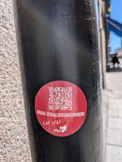 Street sticker A circular, bright red sticker is prominently displayed on a dark, textured metal pole. The upper half of the sticker features a large, white QR code. Below the QR code, in bold white uppercase letters, is the text "RÄDDA SEXUALUNDERVISNINGEN!". Further down, in a more casual, handwritten-style white font, it reads "Låt stå!". In the bottom right quadrant, a white logo is visible, consisting of the letters "rfsu" followed by a stylized white heart symbol. The sticker shows minor signs of wear, suggesting it's been exposed to outdoor elements.