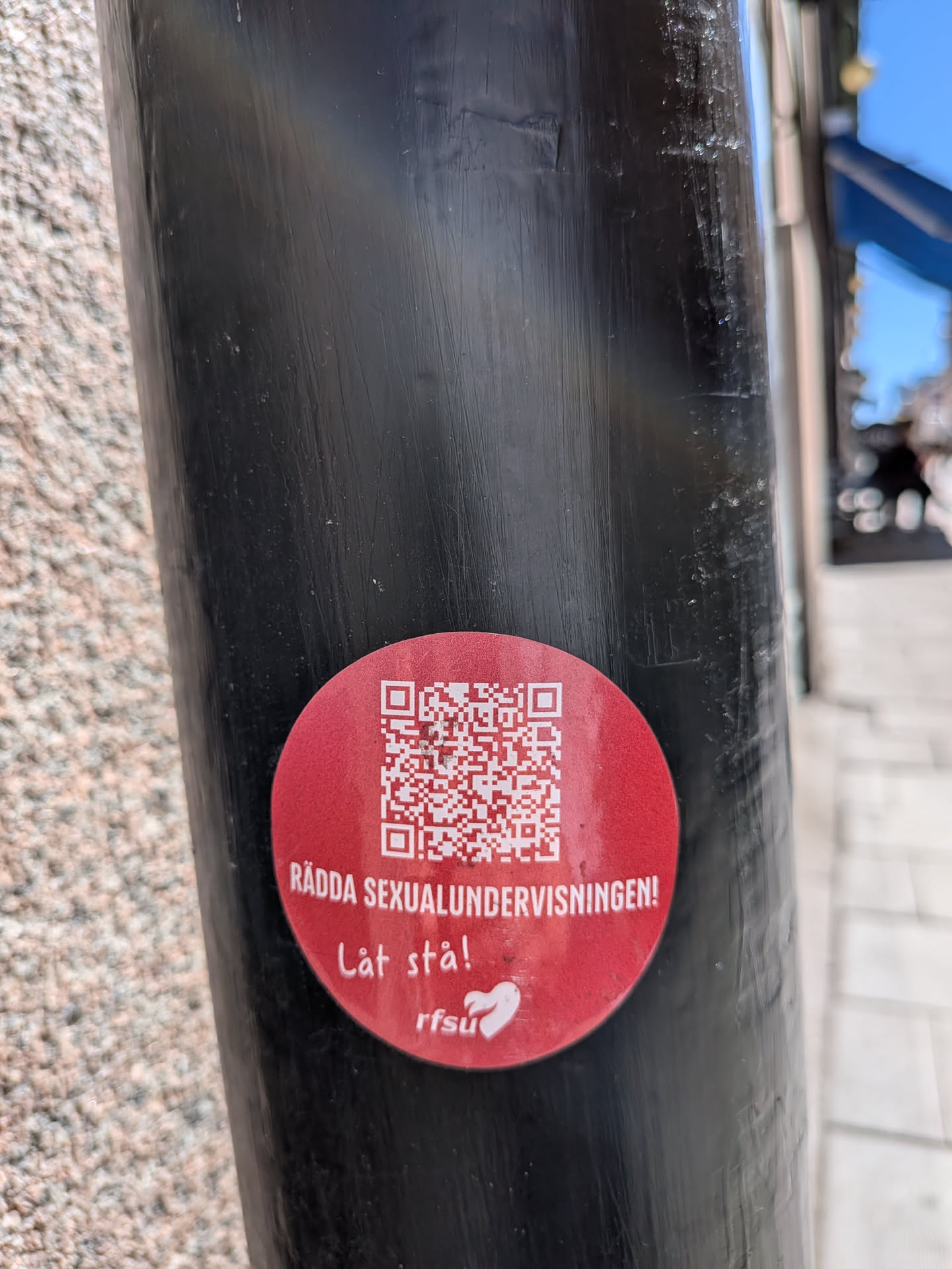 A circular, bright red sticker is prominently displayed on a dark, textured metal pole. The upper half of the sticker features a large, white QR code. Below the QR code, in bold white uppercase letters, is the text "RÄDDA SEXUALUNDERVISNINGEN!". Further down, in a more casual, handwritten-style white font, it reads "Låt stå!". In the bottom right quadrant, a white logo is visible, consisting of the letters "rfsu" followed by a stylized white heart symbol. The sticker shows minor signs of wear, suggesting it's been exposed to outdoor elements.