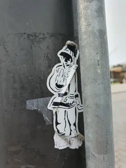 Street sticker A monochrome sticker featuring a stylized figure in uniform, possibly military or historical, making a rude gesture. The sticker is affixed to a metallic pole, showing signs of wear and tear.