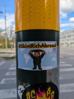 Street sticker This sticker features a cartoon character of a man with a beard wearing a jacket and holding a scarf above his head. The background is a light blue world map. Above the character, the text "@SkintRichAbroad" is written. The sticker also includes the Instagram logo, suggesting a social media presence.