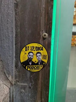 Street sticker A circular yellow sticker affixed to a dark, weathered wooden or metal surface. The sticker features black and white photographic portraits of two men in the center. Above them, the text 'DE OLYDIGA' is printed in a bold, stylized black font. Below them, the word 'PODCAST!' appears in a similar font within a yellow banner. The background is filled with various small black doodles, including a 'Cool S' symbol, stars, an arrow, a heart with an arrow, and crude drawings of male and female anatomy. A QR code is visible on the right side of the sticker.