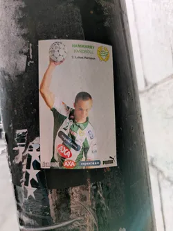 Street sticker The sticker features a picture of Lukas Karlsson, a handball player from Hammarby Handboll, a Swedish handball team. He is holding a handball in his right hand and wearing the team's jersey. The sticker contains logos of sponsors such as Stadium, AXA, expekt.com and Puma.