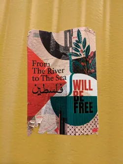 Street sticker A sticker with a design that combines abstract shapes, a plant motif, and text in both English and Arabic. The text on the sticker says, "From The River to The Sea" and "Will Be Free". The sticker shows some damage from being torn and peeling.