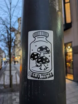 A rectangular, black and white sticker featuring a stylized glass jar with a screw-top lid. Inside the jar, three cartoonish mushrooms with spotted caps and small, root-like feet are depicted. Small circular elements, possibly representing bubbles or spores, are scattered around the mushrooms within the jar. Below the jar, an Instagram logo precedes the text 'BAJO.TINTAS'. The overall style is clean, illustrative, and whimsical.