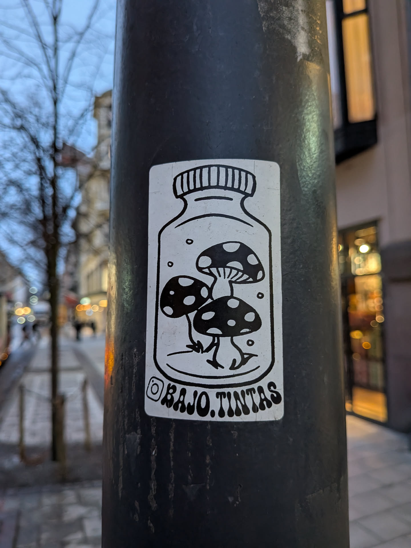 Street sticker A rectangular, black and white sticker featuring a stylized glass jar with a screw-top lid. Inside the jar, three cartoonish mushrooms with spotted caps and small, root-like feet are depicted. Small circular elements, possibly representing bubbles or spores, are scattered around the mushrooms within the jar. Below the jar, an Instagram logo precedes the text 'BAJO.TINTAS'. The overall style is clean, illustrative, and whimsical.