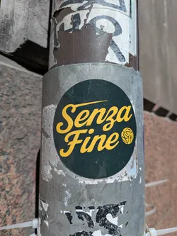 A circular sticker featuring the Italian phrase 'Senza Fine' (meaning 'Without End' or 'Endless') in a vibrant yellow script font against a dark green background. To the right of the text, there is a stylized yellow emblem resembling a ball of yarn or a complex knot. The sticker is applied to a weathered, grey metal pole, with remnants of other peeling stickers visible around its edges.