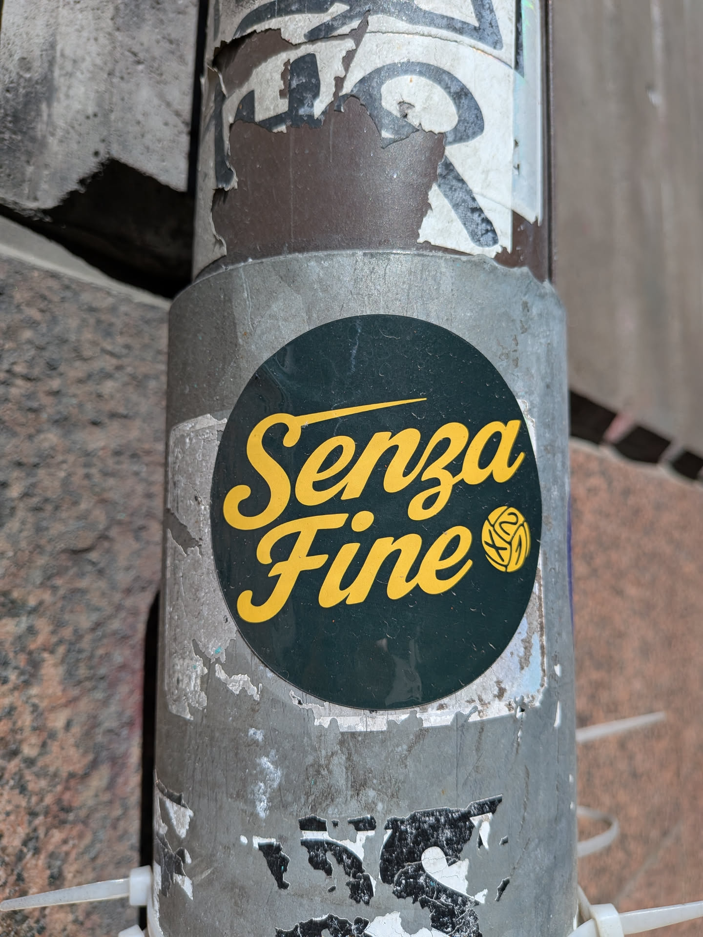 A circular sticker featuring the Italian phrase 'Senza Fine' (meaning 'Without End' or 'Endless') in a vibrant yellow script font against a dark green background. To the right of the text, there is a stylized yellow emblem resembling a ball of yarn or a complex knot. The sticker is applied to a weathered, grey metal pole, with remnants of other peeling stickers visible around its edges.