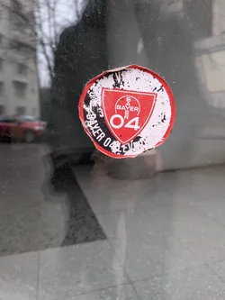 Street sticker A circular sticker featuring the emblem of Bayer 04 Leverkusen. The central design is a red shield with a white outline, containing the word 'BAYER' stacked vertically in white, topped by the Bayer cross logo, and the number '04' in white below it. The shield is set against a black background, which is then surrounded by a white ring. Around the outer edge of the white ring, in black text, are the words 'BAYER 04 LEVERKUSEN VEREIN FÜR LEIBESÜBUNGEN'. The sticker shows significant signs of wear and tear, with frayed edges, peeling, and dirt accumulation, particularly around the black and white outer ring, giving it a distressed and aged appearance.