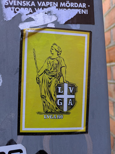 This is a sticker on a wall. It features a yellow background, framed by a white border. The primary image is a drawing of a woman in a classical style, wearing a crown and holding a staff. Next to her is a shield with the letters "LVGA" on it. Below, the word "LVGANO" is printed.