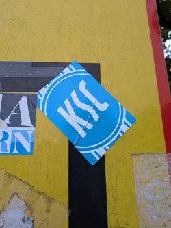 Street sticker A rectangular blue sticker placed diagonally. It features a white circular emblem containing the stylized letters 'KSC' with concentric rings and radial accents.