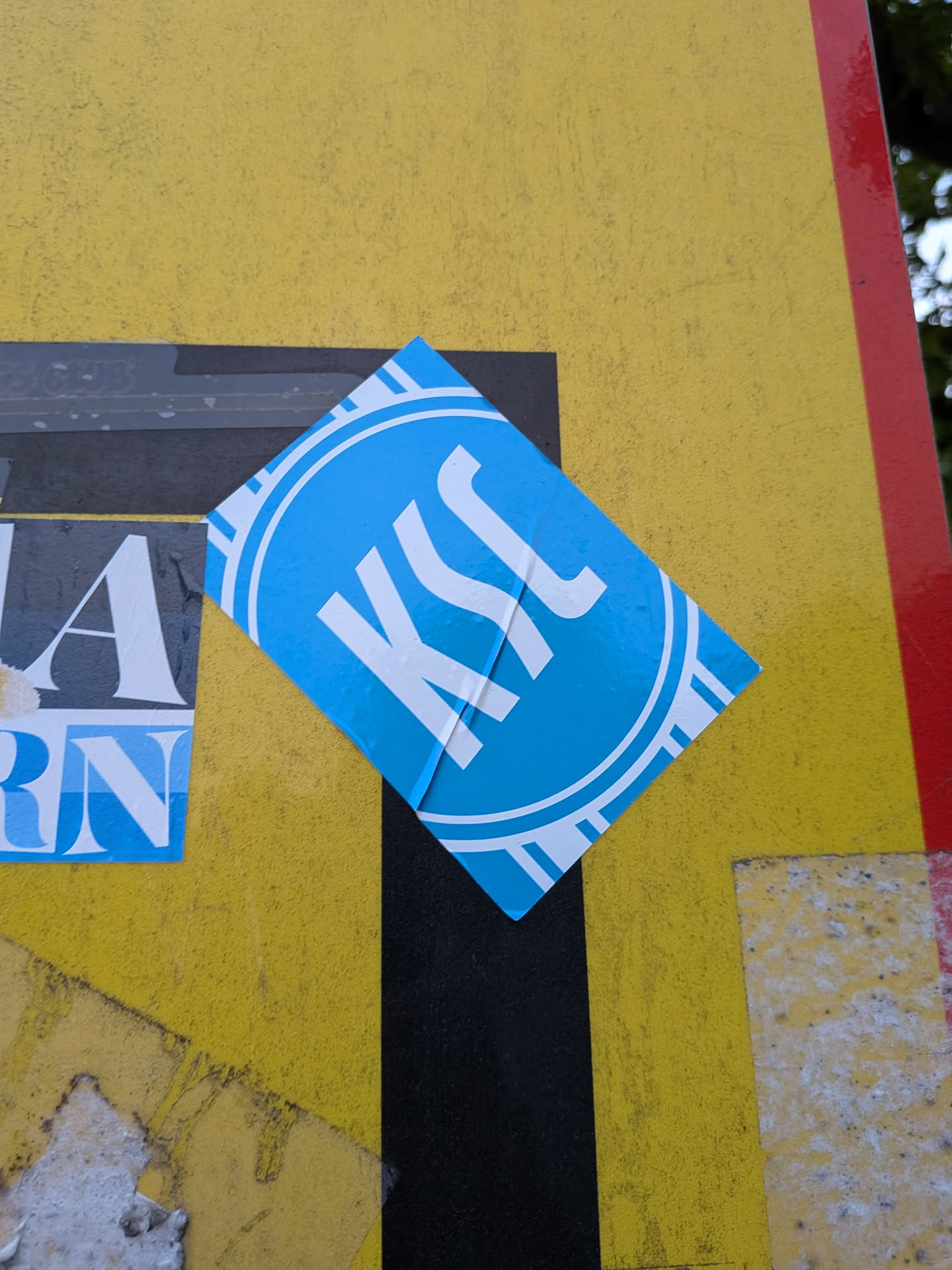 A rectangular blue sticker placed diagonally. It features a white circular emblem containing the stylized letters 'KSC' with concentric rings and radial accents.