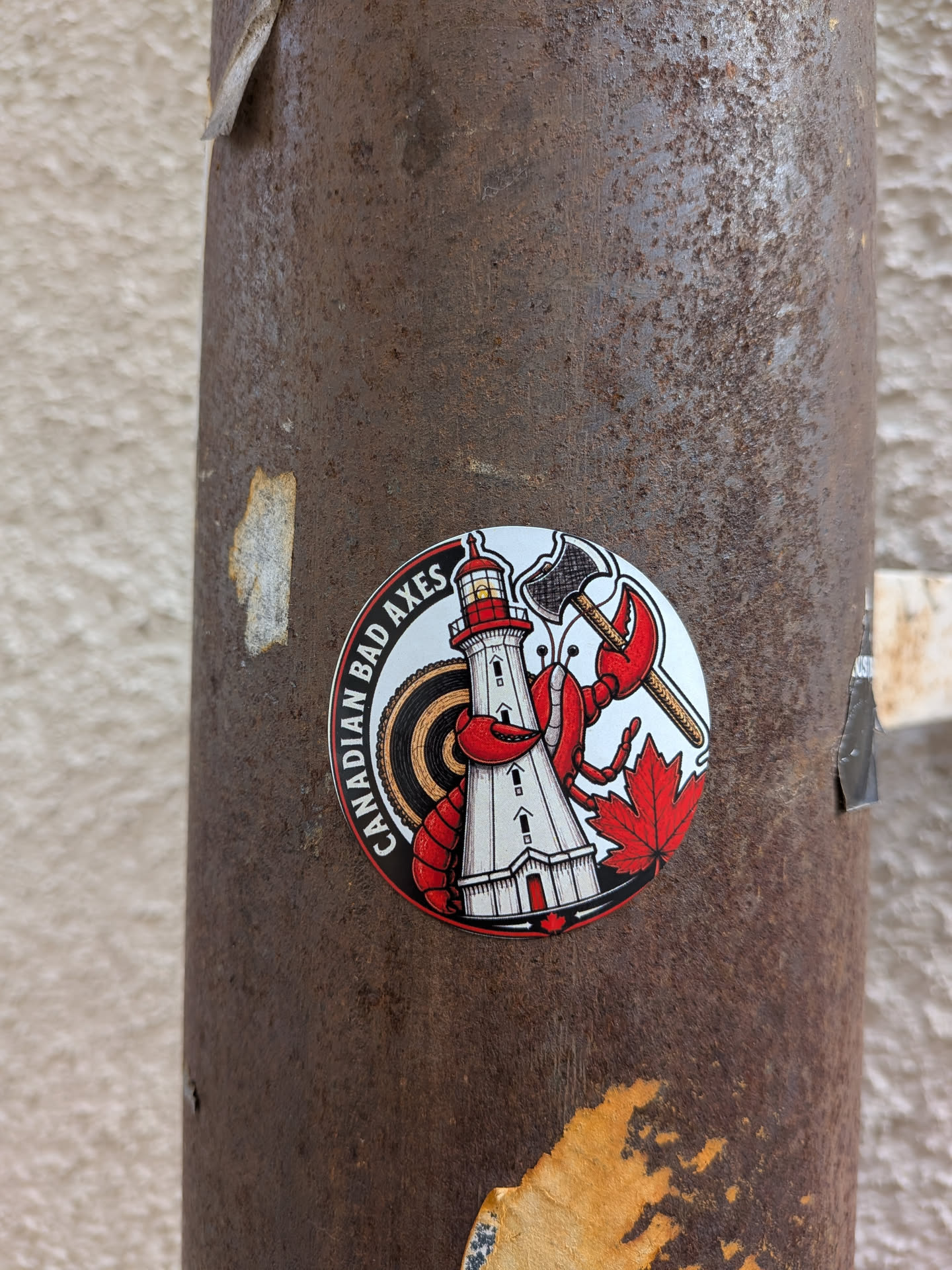 A circular sticker on a rusty metal pole. It depicts a red lobster holding a double-bitted axe next to a white lighthouse, with a target background, a red maple leaf, and the text 'CANADIAN BAD AXES' along the left edge.
