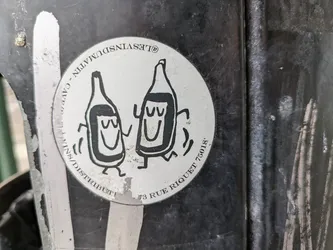 Street sticker A round sticker featuring a simple illustration of two bottles with smiling faces and small legs, seemingly dancing. The text around the image reads 'LES VINS DU MATIN - DISTRIBUTION - 73 RUE RIQUET 75018'. The sticker is adhered to a dark-colored, possibly metallic, surface. The sticker's design is minimal and playful.