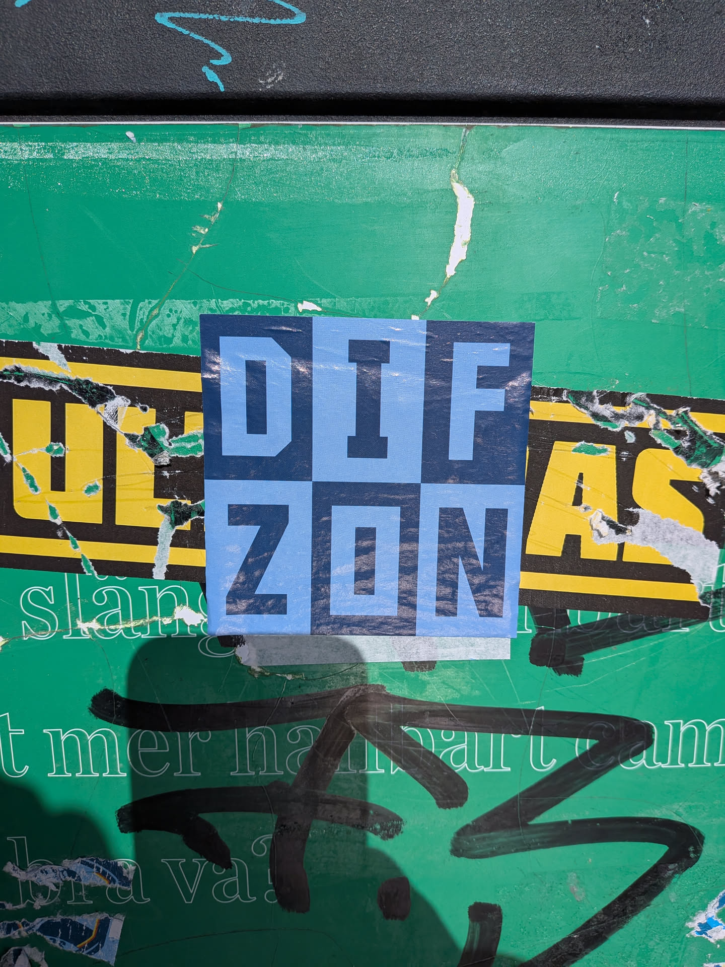 A square sticker featuring a bold, two-tone typographic design. The sticker is divided into a 2x2 grid, with the letters 'D', 'I', 'F' on the top row and 'Z', 'O', 'N' on the bottom row, forming the word 'DIF ZON'. The letters are rendered in a blocky, sans-serif font. The color scheme alternates between a lighter sky blue and a darker navy blue, creating a checkerboard effect where the letters are formed by the contrast. The sticker shows some minor wear and tear, particularly at its bottom edge, and is placed on a green surface covered with other torn posters and graffiti.