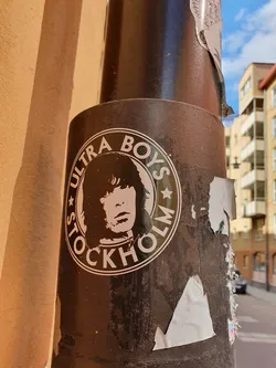 A round sticker featuring a stylized portrait and the text 'Ultra Boys Stockholm'. The sticker is affixed to a dark brown pole outdoors.