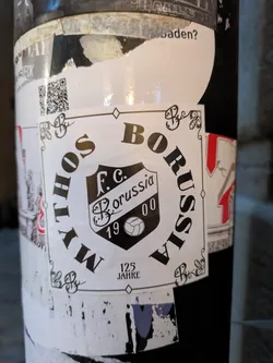 Street sticker The sticker is square-shaped with a white background and a black border. It features ornate black decorative elements in the corners. In the center is a shield-shaped crest with the text 'F.C. Borussia' and '19 00' within it, along with a stylized football. The words 'MYTHOS' and 'BORUSSIA' are printed in large, bold, black capital letters curving around the crest. Below the crest, the text '125 JAHRE' is visible.
