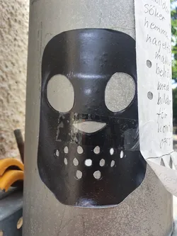 This sticker features a black mask design with cut-out eyes and a small mouth. The mask is reminiscent of a hockey goalie mask or a more anonymous, mysterious persona. The sticker is affixed to a gray pole, with handwritten notes visible nearby.