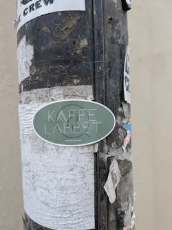 Street sticker An oval-shaped sticker with a muted sage green background and a thin white border. The sticker features the text 'KAFFE LABBET' in a thin, white, stylized sans-serif font. Behind the text is a faint, thin-line circular logo. At the bottom, there is a small Instagram icon followed by the handle 'kaffelabbet.se'.