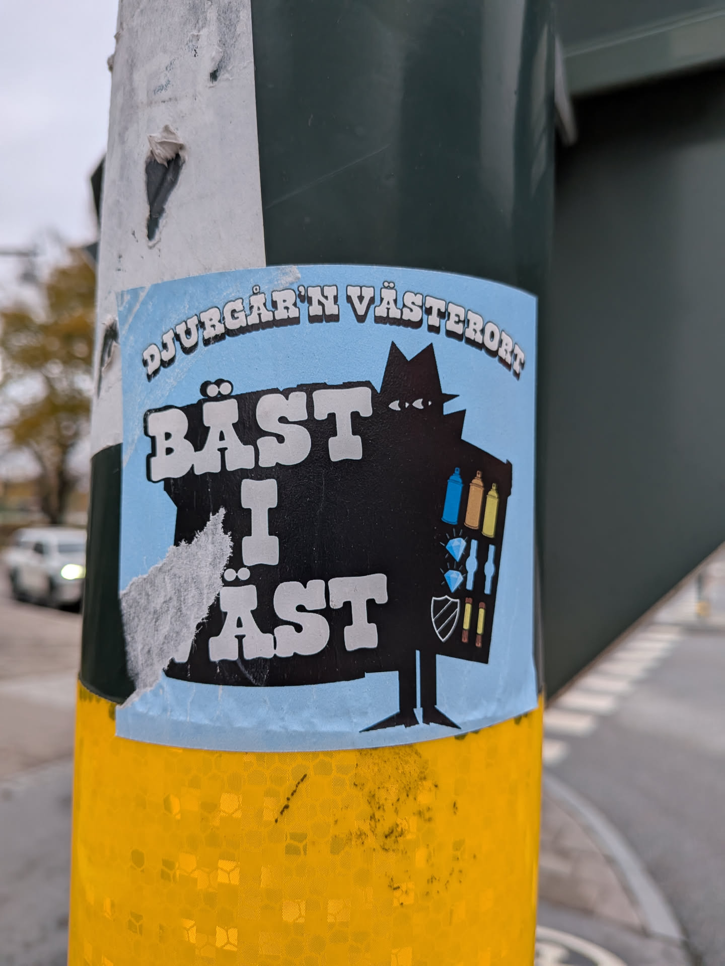 The sticker is predominantly light blue with a black silhouette of a figure wearing a fedora and trench coat. The figure appears to be holding a basket or bag filled with various items. Visible within the silhouette are three spray paint cans (blue, gold, yellow), two diamond shapes, a shield emblem, and two more spray paint cans (red and yellow). The text 'DJURGÅR'N VÄSTERORT' is arched across the top in white, outlined in black. Below this, in large, bold white letters with a black outline, is the text 'BÄST I ÄST'. The sticker shows signs of wear and tear, with some peeling and creasing, particularly on the left side.
