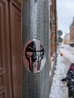The sticker is oval-shaped and features a highly stylized, metallic-looking mask, strongly resembling the iconic mask of rapper MF DOOM. The design is rendered in black and reflective silver/chrome, with the silver areas catching ambient light and showing subtle color reflections from the surroundings (e.g., hints of blue and orange/brown). The mask has prominent dark, angular eye slits, a defined nose bridge, and a segmented lower face/jawline. A small, diamond-shaped detail with red and blue segments is visible on the upper left side of the mask's forehead. The overall appearance is sharp, geometric, and iconic.