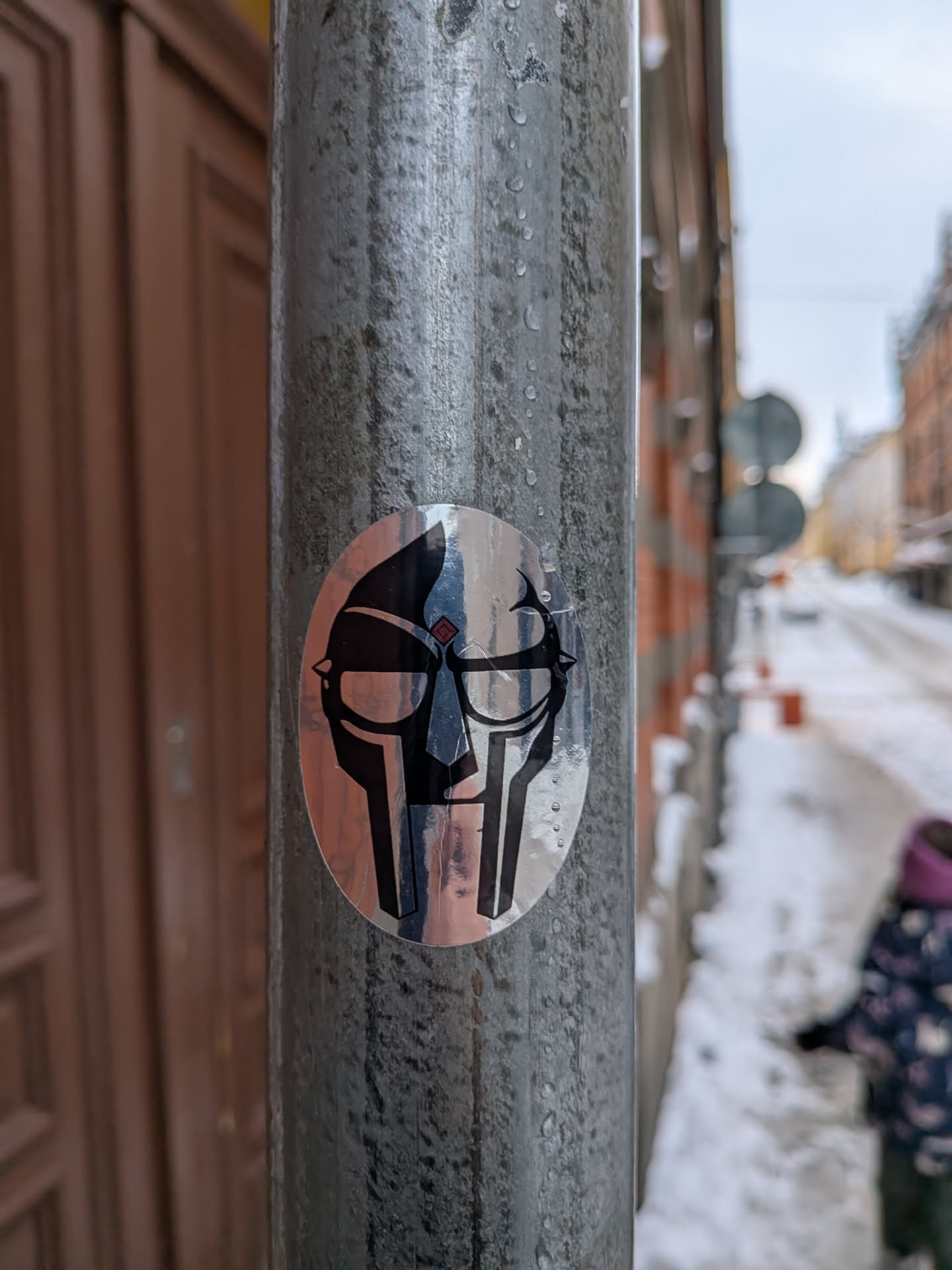 Street sticker The sticker is oval-shaped and features a highly stylized, metallic-looking mask, strongly resembling the iconic mask of rapper MF DOOM. The design is rendered in black and reflective silver/chrome, with the silver areas catching ambient light and showing subtle color reflections from the surroundings (e.g., hints of blue and orange/brown). The mask has prominent dark, angular eye slits, a defined nose bridge, and a segmented lower face/jawline. A small, diamond-shaped detail with red and blue segments is visible on the upper left side of the mask's forehead. The overall appearance is sharp, geometric, and iconic.