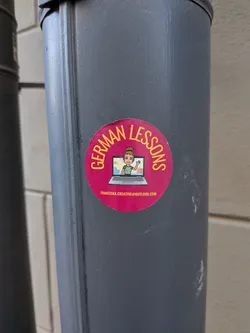 Street sticker A circular sticker with a vibrant magenta background. The top half features the words 'GERMAN LESSONS' in yellow capital letters, curving along the edge. In the center, there's an illustration of a silver laptop with a screen displaying a cartoon avatar of a woman. The avatar has light brown hair in a bun, green eyes, and is wearing a green top, waving with her right hand. Below the laptop, in smaller white text, an email address is visible.