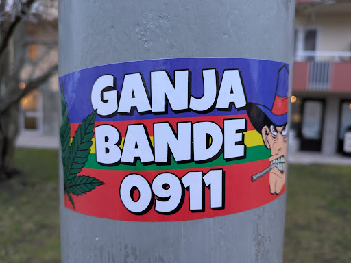 Street sticker A sticker featuring a cartoon illustration of a man in a hat smoking a cigarette, with the text 'GANJA BANDE 0911' and a marijuana leaf. The background is a colorful horizontal stripe pattern.