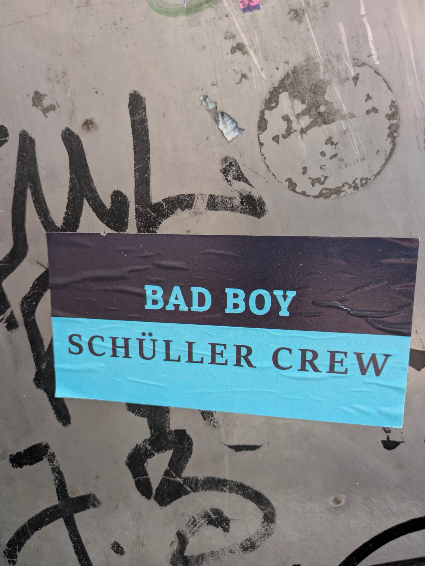 A horizontal rectangular sticker adhered to a gray, grungy surface with black graffiti tags. The sticker is divided into two horizontal bands: the top band is a dark blue or black, and the bottom band is a bright light blue or turquoise. The words "BAD BOY" are prominently displayed in light blue text on the dark upper band, and "SCHÜLLER CREW" is written in dark text on the light blue lower band. The sticker shows some signs of wear and slight wrinkling.