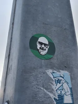 Street sticker A round sticker featuring a portrait of a bald man wearing sunglasses. The portrait is in black and white, and the background is green with horizontal stripes.  There's a second, partially torn sticker below it featuring a figure in a hat and coat.