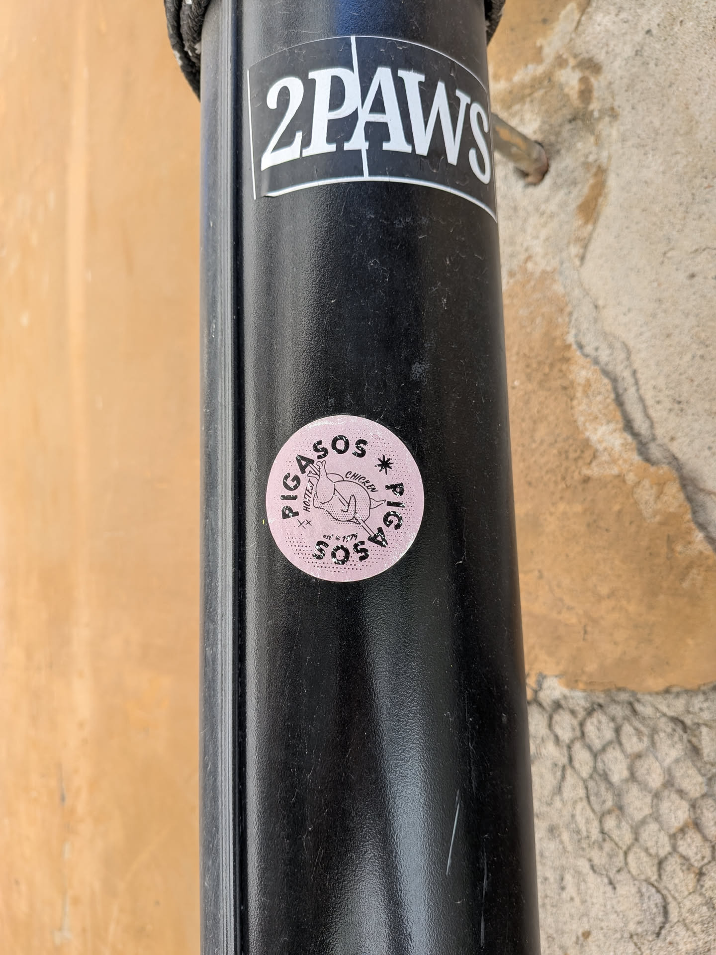 A circular sticker featuring a light pink background with a subtle dotted texture. The central graphic is a stylized, cartoonish chicken or chicken wing, rendered in black outline, depicted with small wings as if in flight or falling. Text is arranged around and below the central image. 'PIGASOS' and 'CHICKEN' are curved along the top, while 'HOTEST' and 'SOS' are positioned below the chicken. A small 'est * 2019' is at the very bottom. The design also incorporates small stars and cross-hatch patterns, giving it a slightly distressed or vintage aesthetic. The overall color palette is pink and black.