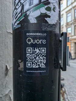 A rectangular black sticker featuring white text and a yellow-gold logo. At the top, 'BORGOBELLO' is written in white uppercase letters. Below it, the word 'Quore' is prominently displayed in a stylized yellow-gold font, with the 'Q' forming a heart-like shape. A large, white QR code is centered below the logo. At the very bottom, 'quoreimmobiliare.it' is written in smaller white text. The sticker is applied to a dark pole and shows some minor dust and wear, indicating it's a real-world application.