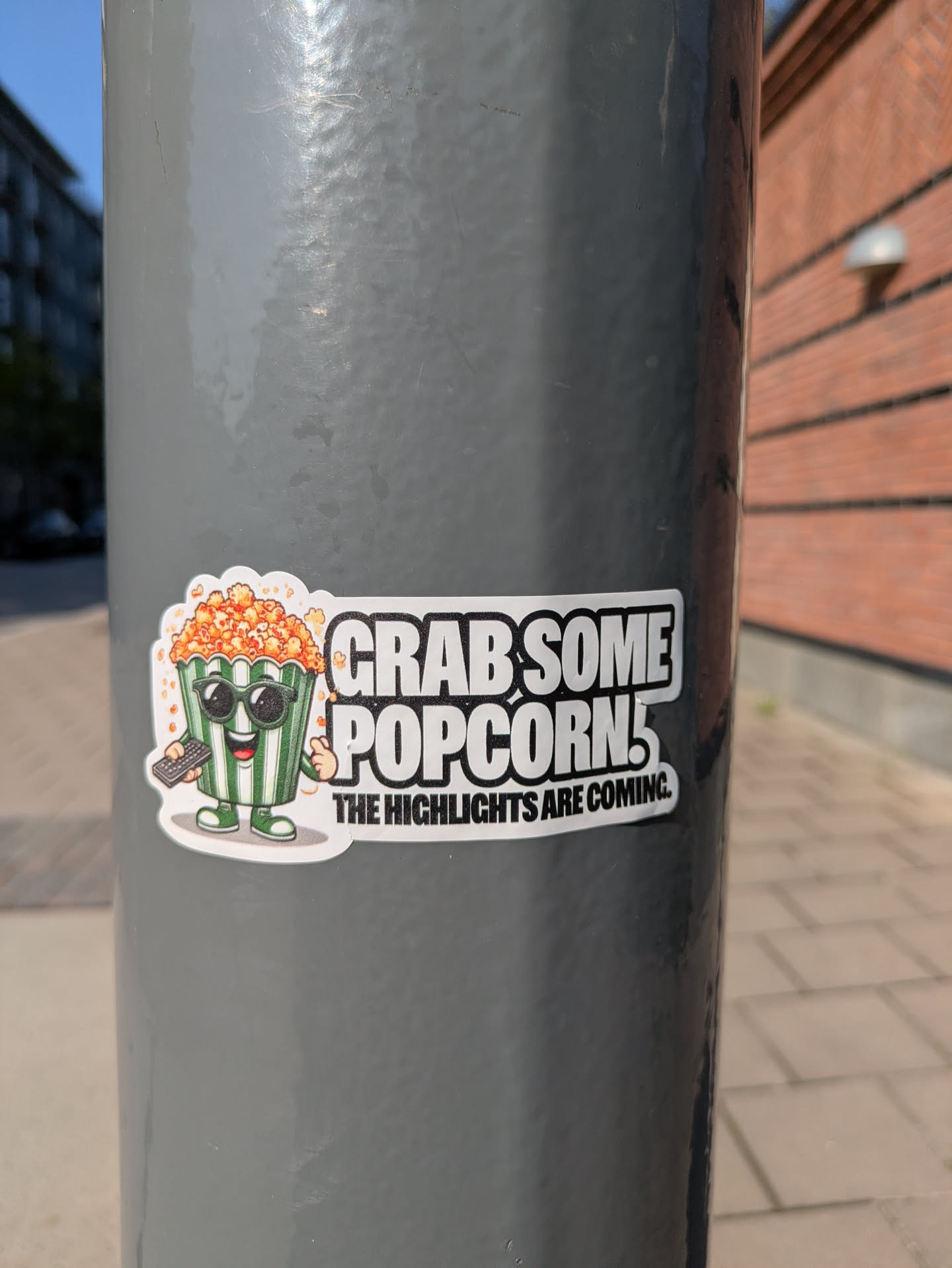 This sticker depicts a popcorn container with sunglasses, personified with arms, legs and a smiling face. The container has green and white stripes and is full of popcorn. It holds a remote control in one hand. The sticker also includes the text "Grab some popcorn! The highlights are coming."