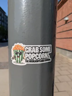 Street sticker This sticker depicts a popcorn container with sunglasses, personified with arms, legs and a smiling face. The container has green and white stripes and is full of popcorn. It holds a remote control in one hand. The sticker also includes the text "Grab some popcorn! The highlights are coming."