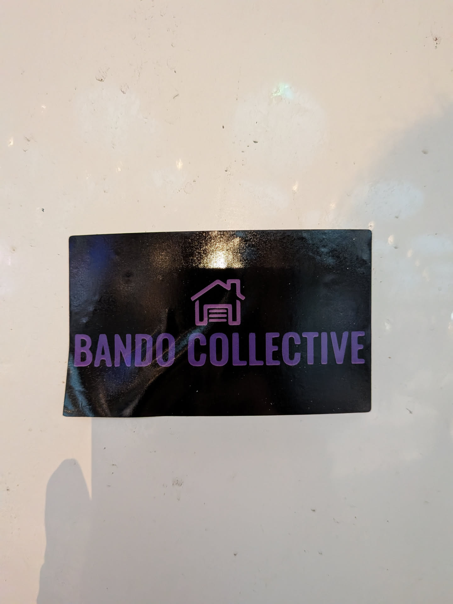 Street sticker The sticker is rectangular with rounded corners and a glossy black background. Centered on the sticker is a stylized purple icon depicting a house with a garage door. To the right of the icon, the text 'BANDO COLLECTIVE' is written in a bold, sans-serif purple font. The overall aesthetic is clean and modern.