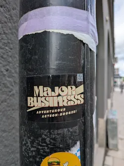 A rectangular sticker with a black background featuring bold, cream-colored text that reads 'Major Business'. The letters have a slight orange-brown shadow, giving them a 3D, retro appearance. A light yellow diagonal stripe or banner runs behind and through the word 'Business'. Below the main text, in smaller, cream-colored sans-serif font, it says 'ADVENTUROUS ACTION-BOOGIE!'. In the top right corner, there is a small, black and white QR code.