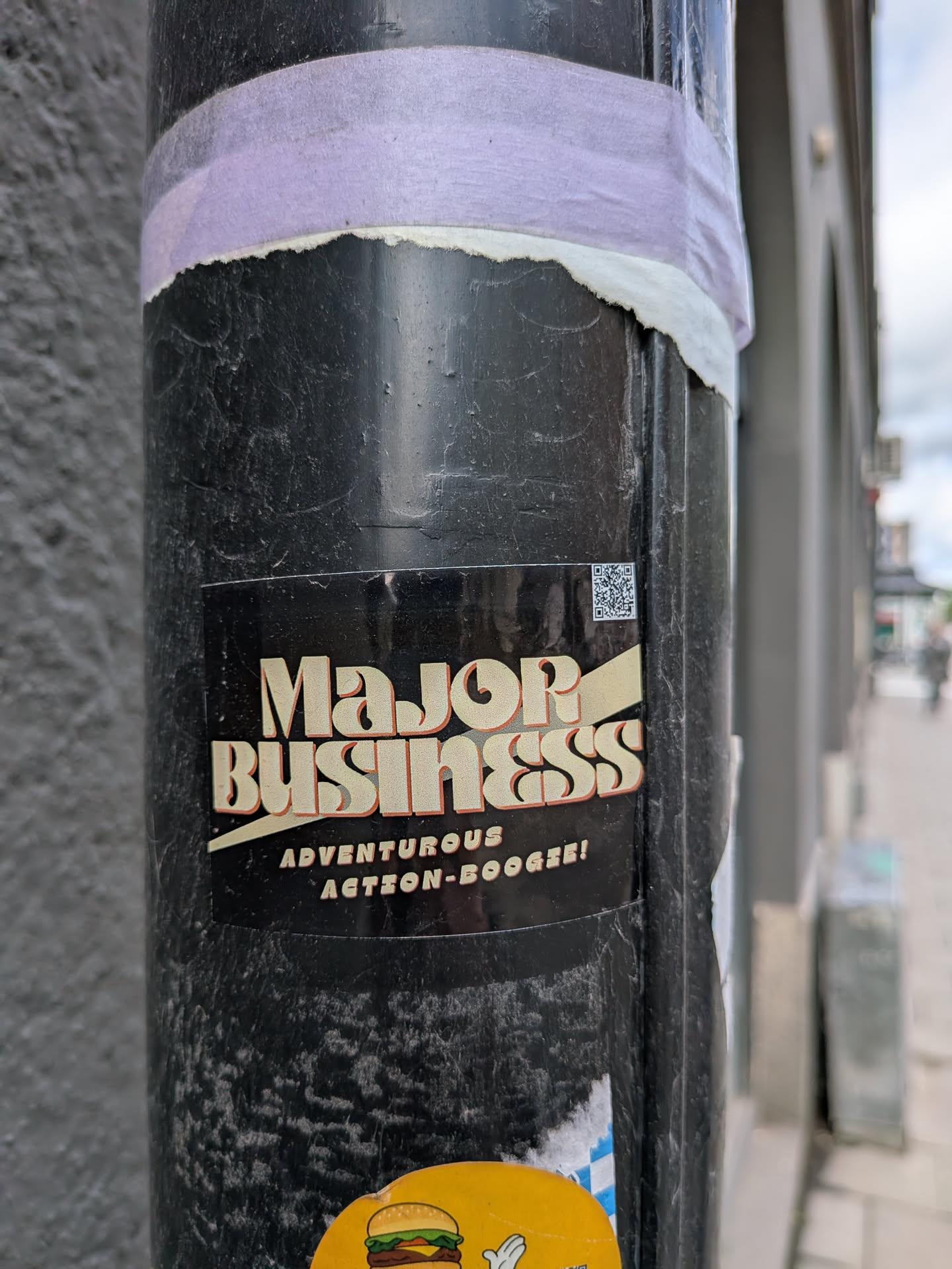 A rectangular sticker with a black background featuring bold, cream-colored text that reads 'Major Business'. The letters have a slight orange-brown shadow, giving them a 3D, retro appearance. A light yellow diagonal stripe or banner runs behind and through the word 'Business'. Below the main text, in smaller, cream-colored sans-serif font, it says 'ADVENTUROUS ACTION-BOOGIE!'. In the top right corner, there is a small, black and white QR code.