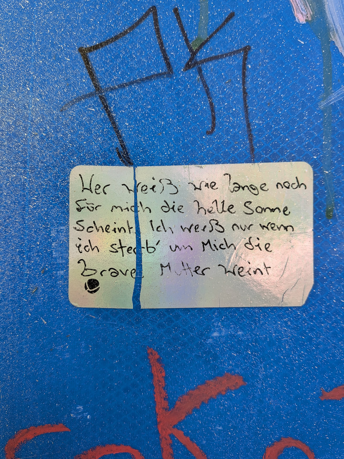 A rectangular sticker with rounded corners, featuring a slightly iridescent or holographic surface, is adhered to a textured, speckled blue background. The sticker is split by a prominent vertical tear or crease down its center. Black handwritten German text covers the sticker, appearing in a neat, cursive-like script. A small, stylized black symbol (resembling an 'e' or a simple logo) is visible in the bottom-left corner. The sticker's edges show minor wear, and it is surrounded by other graffiti elements on the blue surface.