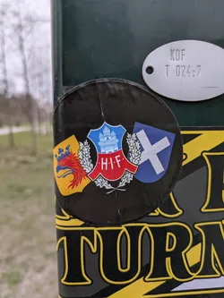 Street sticker The sticker shows three shields, each with different designs and colors. The central shield features a blue and white design with a crown and buildings, surrounded by laurel. To the left is a shield with a red griffin on a yellow background, and to the right, a blue shield with a white saltire. The letters 'H.I.F.' are prominently displayed on the central shield.