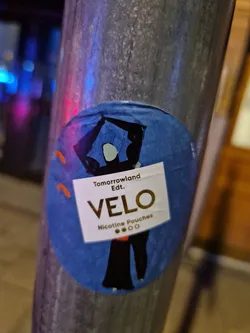 A round sticker featuring a stylized illustration of two figures under a house-like structure.  The text 'VELO' is prominently displayed with the tagline 'Nicotine Pouches' and 'Tomorrowland Edt.'