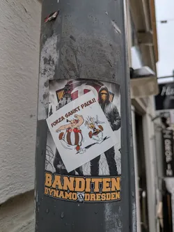 The central image features two stickers layered on a weathered grey pole. The larger, background sticker has a dark, somewhat gritty aesthetic, depicting a hooded figure with a face covering, standing amidst barbed wire. In the upper background, a stylized representation of the Frauenkirche in Dresden is visible. Below the figure, prominent orange-yellow text reads "BANDITEN" above "DYNAMO DRESDEN", with the Dynamo Dresden club crest integrated. Overlaid on this, slightly askew, is a smaller, white sticker featuring the classic cartoon characters Obelix and Asterix. Obelix is shown flexing his arm, while Asterix runs alongside him. Above them, brown text declares "FORZA SANKT PAULI!". Both stickers show signs of outdoor exposure, including water droplets.