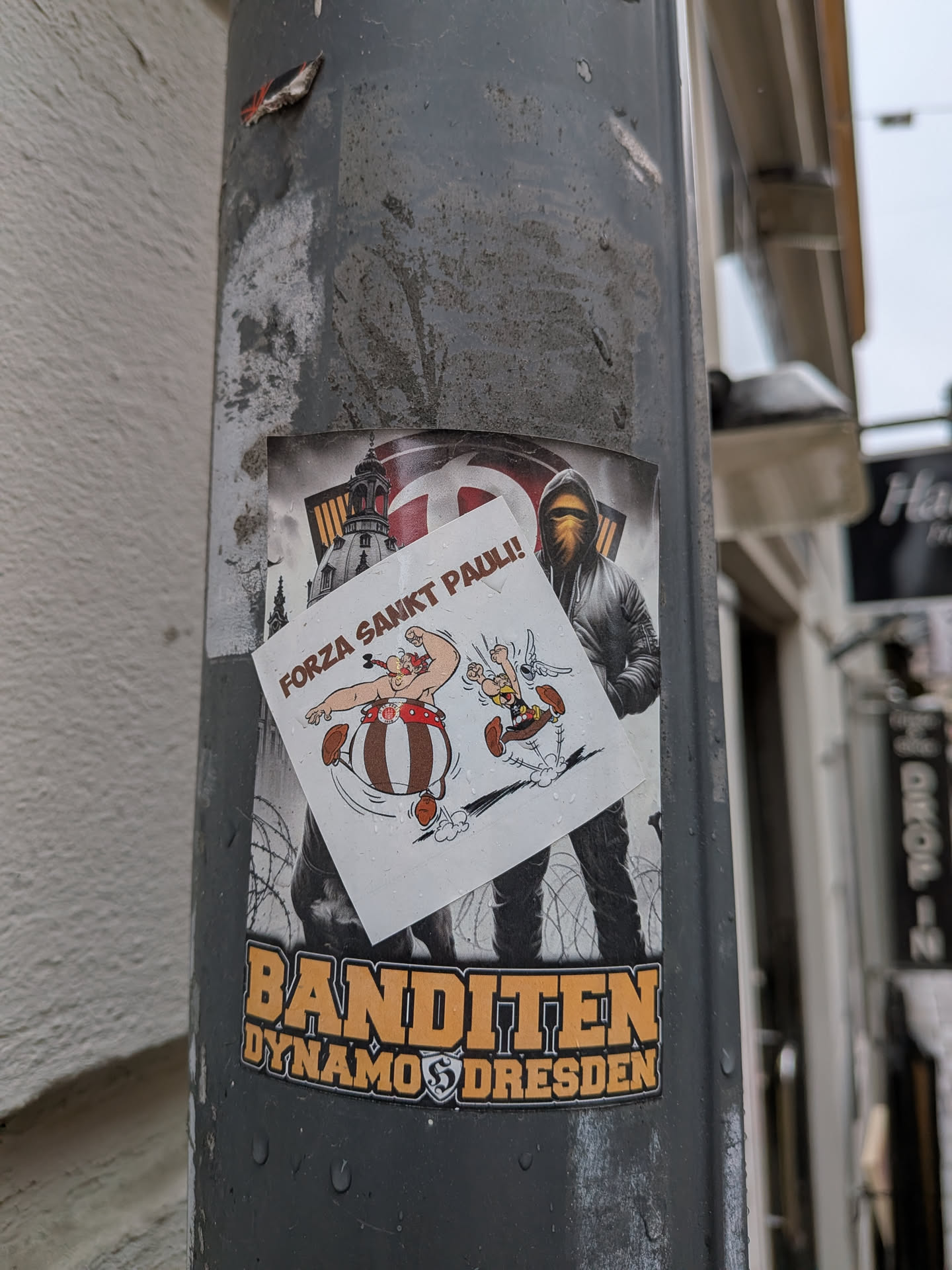 The central image features two stickers layered on a weathered grey pole. The larger, background sticker has a dark, somewhat gritty aesthetic, depicting a hooded figure with a face covering, standing amidst barbed wire. In the upper background, a stylized representation of the Frauenkirche in Dresden is visible. Below the figure, prominent orange-yellow text reads "BANDITEN" above "DYNAMO DRESDEN", with the Dynamo Dresden club crest integrated. Overlaid on this, slightly askew, is a smaller, white sticker featuring the classic cartoon characters Obelix and Asterix. Obelix is shown flexing his arm, while Asterix runs alongside him. Above them, brown text declares "FORZA SANKT PAULI!". Both stickers show signs of outdoor exposure, including water droplets.