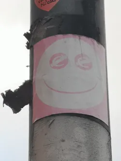 Street sticker The sticker features a large, white, oval-shaped face with a pink outline, set against a pink background. The eyes are two pink circles, each containing a stylized white 'e' or 'c' symbol. A simple, curved pink line forms a wide, friendly smile. The sticker is affixed to a dark, textured pole and shows signs of wear, including peeling edges and some dirt accumulation, particularly on the pole itself.