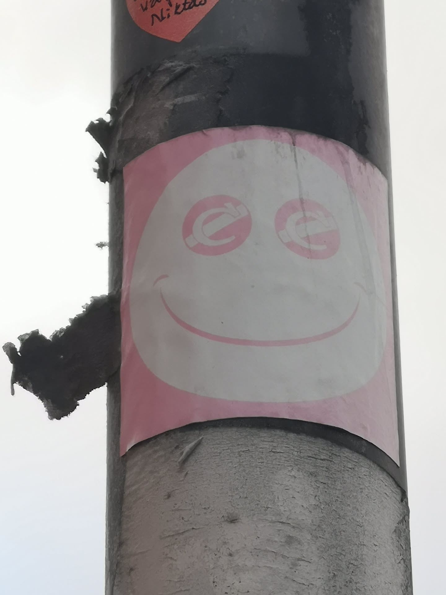 The sticker features a large, white, oval-shaped face with a pink outline, set against a pink background. The eyes are two pink circles, each containing a stylized white 'e' or 'c' symbol. A simple, curved pink line forms a wide, friendly smile. The sticker is affixed to a dark, textured pole and shows signs of wear, including peeling edges and some dirt accumulation, particularly on the pole itself.