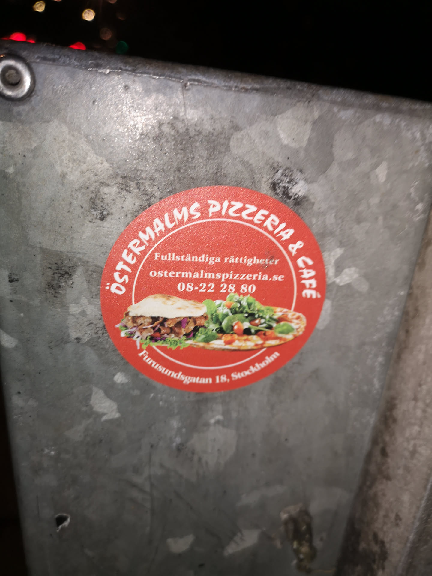 A vibrant circular red sticker with white text and two appetizing food images. The top arc of the sticker reads 'ÖSTERMALMS PIZZERIA & CAFÉ', while the bottom arc displays the address 'Furusundsgatan 18, Stockholm'. Inside the circle, there's text 'Fullständiga rättigheter', the website 'ostermalmspizzeria.se', and a phone number '08-22 28 80'. The central imagery features a hearty kebab in pita bread with fresh vegetables on the left, and a pizza topped with green leaves (likely arugula) and cherry tomatoes on the right, all set against the bright red background.