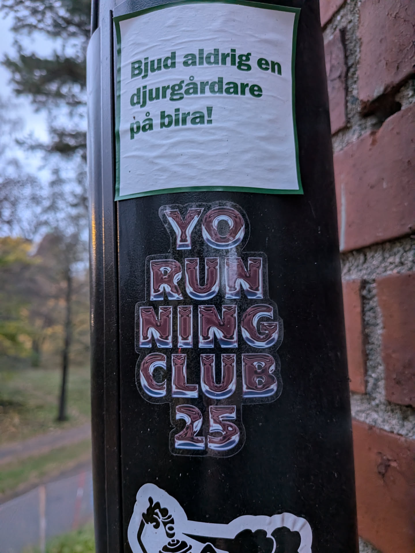 The sticker is centrally located on a dark, cylindrical surface, likely a lamppost. It features bold, stylized text in a metallic, reflective font with a dark red fill and a white outline. The text reads 'YO RUNNING CLUB 25' stacked vertically. The letters have a glossy, almost chrome-like appearance, with subtle shading that gives them a 3D effect. Below this text, a portion of another white sticker with a black outline is visible, depicting a stylized figure or emblem.