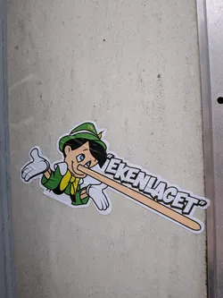 Street sticker The sticker features a cartoon character resembling Pinocchio, wearing a green hat with a yellow emblem, a green jacket, and a yellow scarf. His nose is exaggeratedly long and extends horizontally across the sticker, transforming into text. The character has blue eyes, black hair, and is depicted with white gloves on both hands, one of which is raised in a gesture. The overall color scheme is vibrant, with green, yellow, white, and black being prominent. The text is in bold, white, block letters with black outlines, appearing to be part of the extended nose.