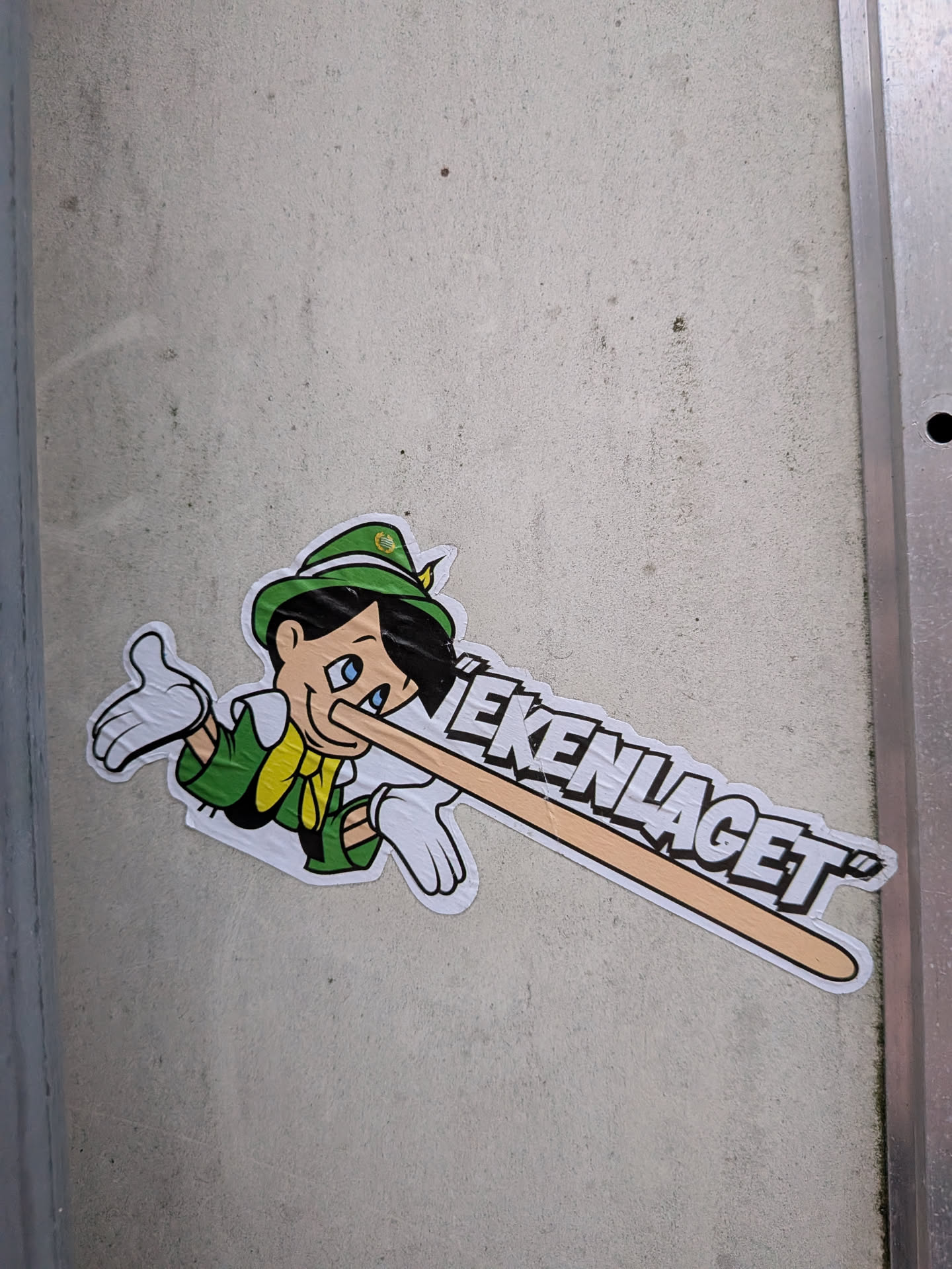 Street sticker The sticker features a cartoon character resembling Pinocchio, wearing a green hat with a yellow emblem, a green jacket, and a yellow scarf. His nose is exaggeratedly long and extends horizontally across the sticker, transforming into text. The character has blue eyes, black hair, and is depicted with white gloves on both hands, one of which is raised in a gesture. The overall color scheme is vibrant, with green, yellow, white, and black being prominent. The text is in bold, white, block letters with black outlines, appearing to be part of the extended nose.
